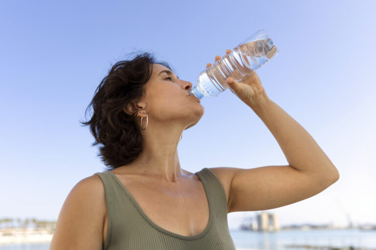 Mujer hidratándose al aire libre con agua embotellada en un día soleado, ideal para ilustrar temas sobre consumo diario de agua y salud