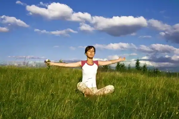 Mujer sentada en posición de meditación en un campo verde, con los brazos extendidos y los ojos cerrados, practicando técnicas de relajación al aire libre bajo un cielo azul con nubes.