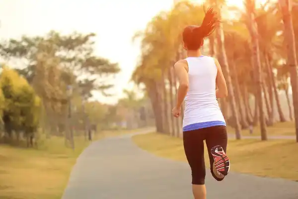Mujer corriendo por un sendero al aire libre al atardecer, rodeada de árboles, como parte de una rutina de ejercicio físico para reducir el estrés.