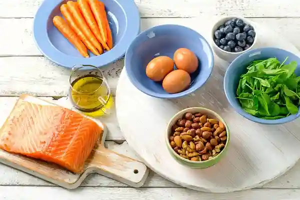 Alimentos saludables sobre una mesa de madera: salmón, zanahorias, huevos, espinaca, arándanos, frutos secos y aceite de oliva, ideales para una dieta equilibrada.