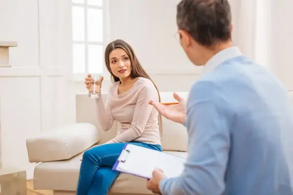 Mujer joven sentada en un sofá conversando con un terapeuta mientras sostiene un vaso de agua, durante una sesión en un entorno luminoso y tranquilo.