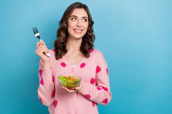 Mujer joven con suéter rosa de corazones sosteniendo un bowl de ensalada y un tenedor, sonriendo y mirando hacia un lado sobre fondo azul.